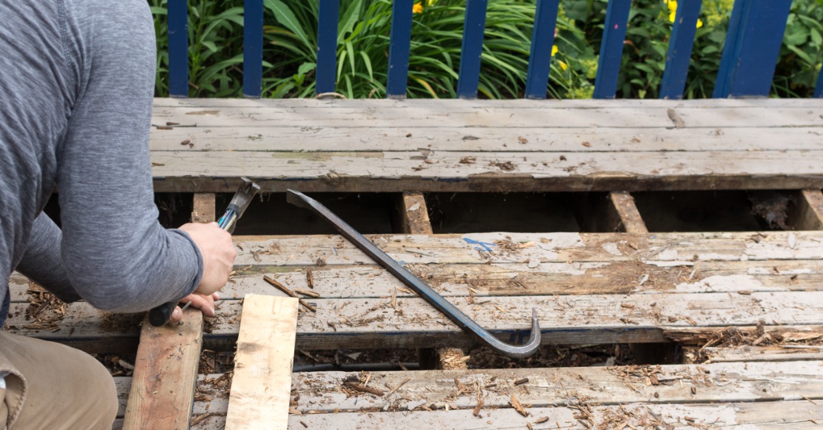 A close-up of a man using a hammer and a metal bar to remove old wood from the base of a deck with a metal rail.